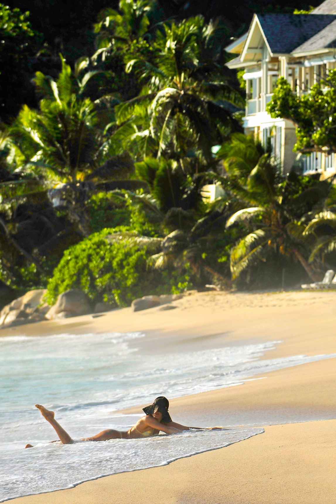 A woman lying in the shallow surf below a tropical resort — the landscape a guest wants to enter.
