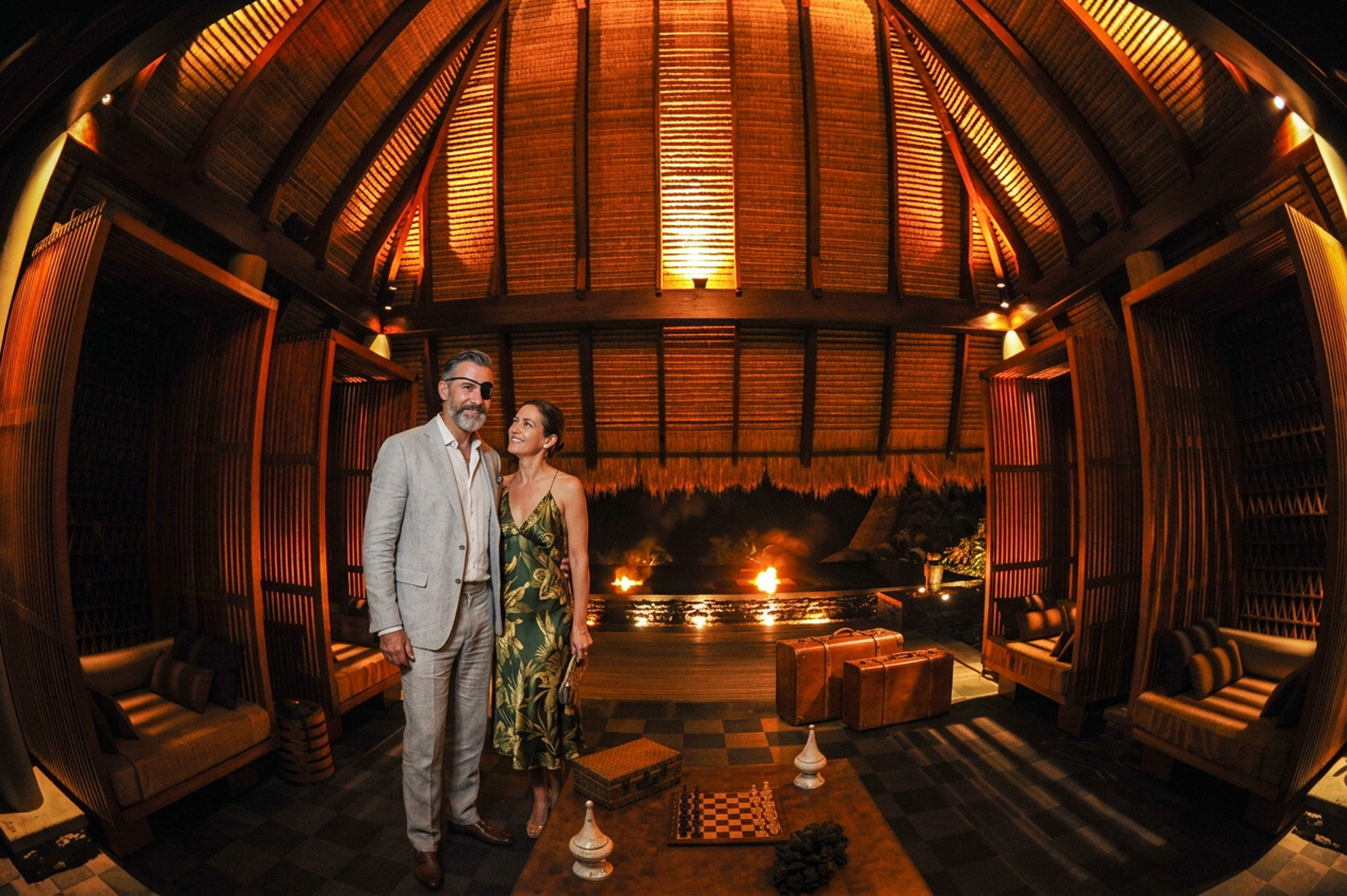 A couple standing under the lit timber dome of a Seychelles resort pavilion, fires burning at the water beyond — the moment a stay is imagined rather than described.