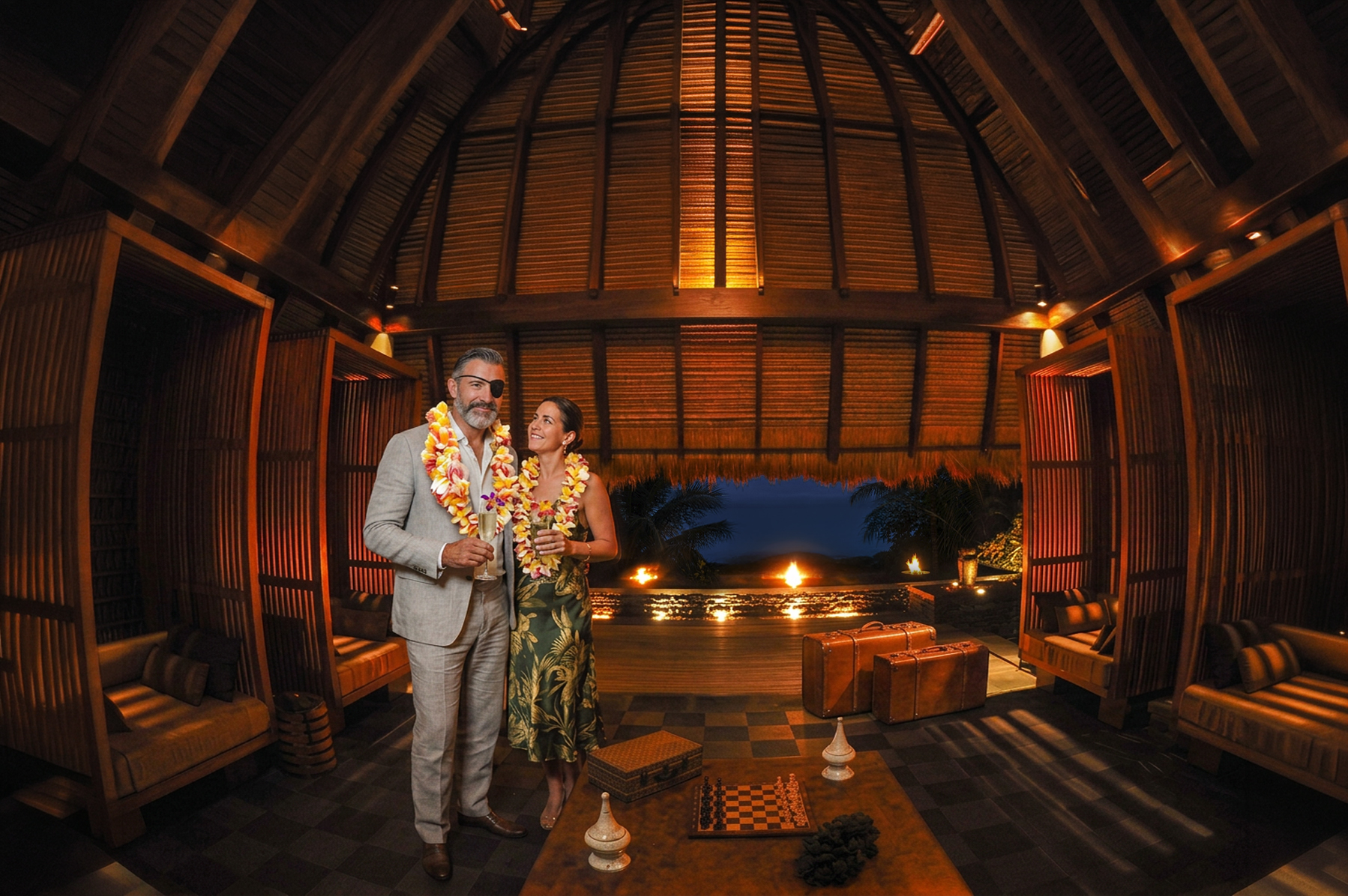 A couple standing under the lit timber dome of a Seychelles resort pavilion, fires burning at the water beyond — the moment a stay is imagined rather than described.